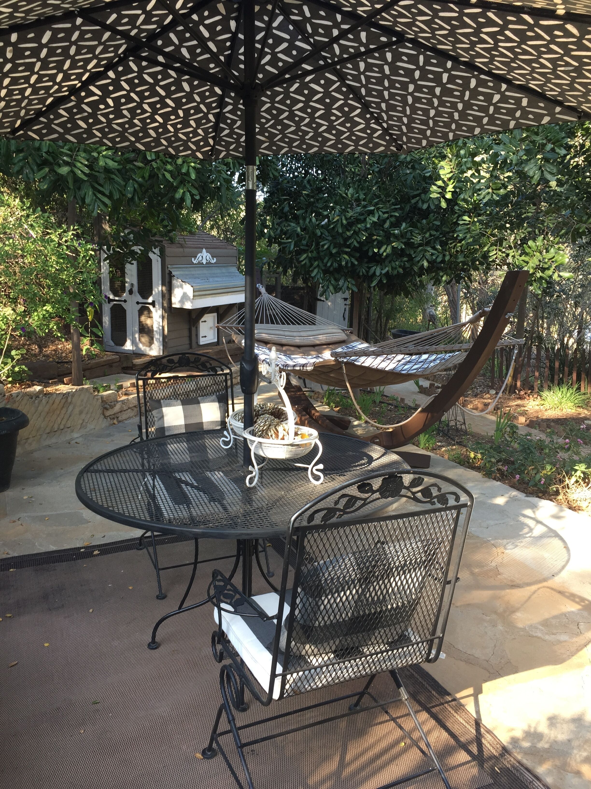 Een terras buiten met een ronde metalen tafel en stoelen, een hangmat en een barbecueplek.