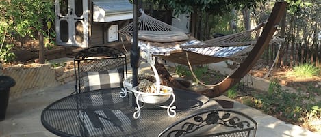 Een terras buiten met een ronde metalen tafel en stoelen, een hangmat en een barbecueplek.