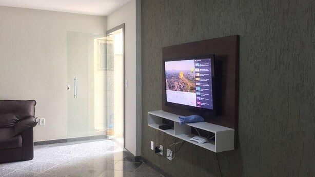 A modern living room with a flat-screen TV mounted on a wall, a leather sofa, and a glass door leading to another room. - Condomínio Ed. Sirene Fraga (Guarapari)