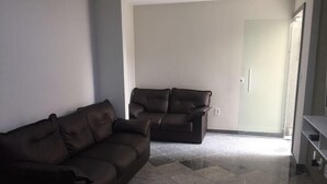 A living room with a brown sofa and a recliner, tiled flooring, and a door leading to another room. - Condomínio Ed. Sirene Fraga (Guarapari)