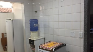 A kitchen with white tiled walls, a white refrigerator, a blue water dispenser, and a stove with a colorful cloth. - Condomínio Ed. Sirene Fraga (Guarapari)