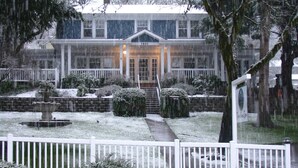 A snow-covered garden with a fountain, a white picket fence, and a house with a number sign.