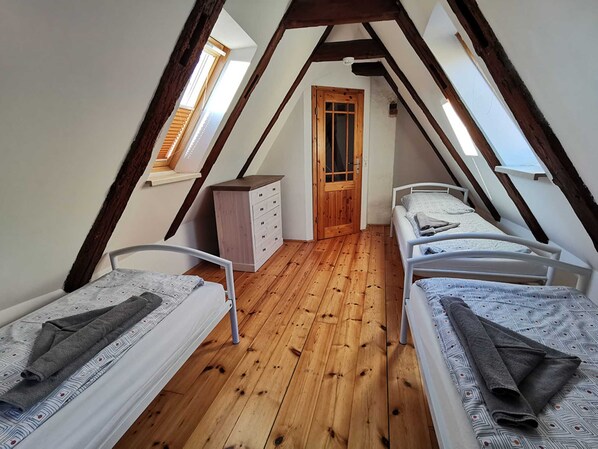 Un pequeño dormitorio en el ático con dos camas individuales, suelo de madera, un armario de madera y una puerta que da a otra habitación.