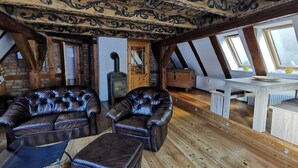 Ein Schlafzimmer im Loft-Stil mit schrägem Dach, Holzbalken, Kamin, zwei Ledersesseln, einem kleinen Tisch und einem Fenster mit Blick.