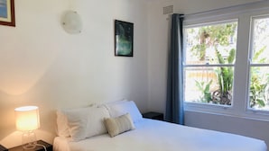 Un dormitorio con cama, lámpara de noche, una obra de arte enmarcada y una ventana con cortinas.