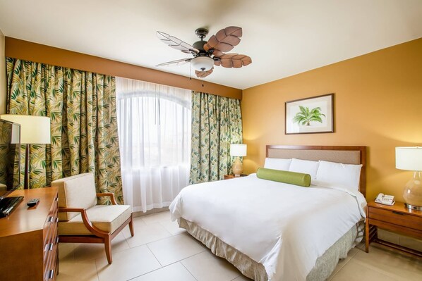 Habitación de hotel con cama, escritorio, silla, ventilador de techo y un cuadro en la pared.