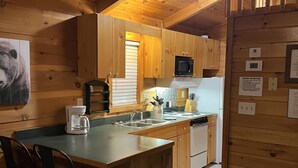 Una cocina acogedora de estilo cabaña con armarios de madera, una isla central con fregadero, microondas y refrigerador. Hay dos taburetes de bar y una mesa de comedor.