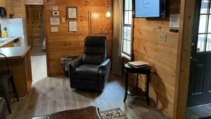Interior acogedor de una cabaña de madera con comedor, un sillón de cuero negro y un televisor con el logotipo de la Fundación para el Liderazgo en el Reciclaje.