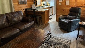 Interior acogedor de una cabaña con sofá y sillón de cuero, una mesa de comedor de madera y una zona de cocina con refrigerador, microondas y lavabo.