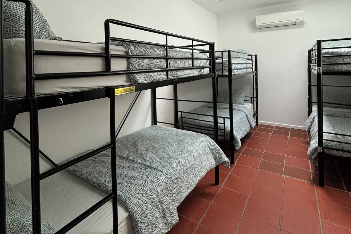 Une chambre de dortoir avec des lits superposés, un sol carrelé, un luminaire au plafond et un appareil de climatisation.