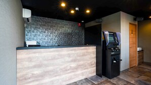 A reception area with a wooden reception desk, a digital kiosk, and a security camera on the wall. - HOTEL R9 The Yard Ashikagafukutomi (Ashikaga)