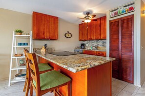 A kitchen with wooden cabinets, a granite countertop, a ceiling fan, a clock, and a shelf with decorative items.