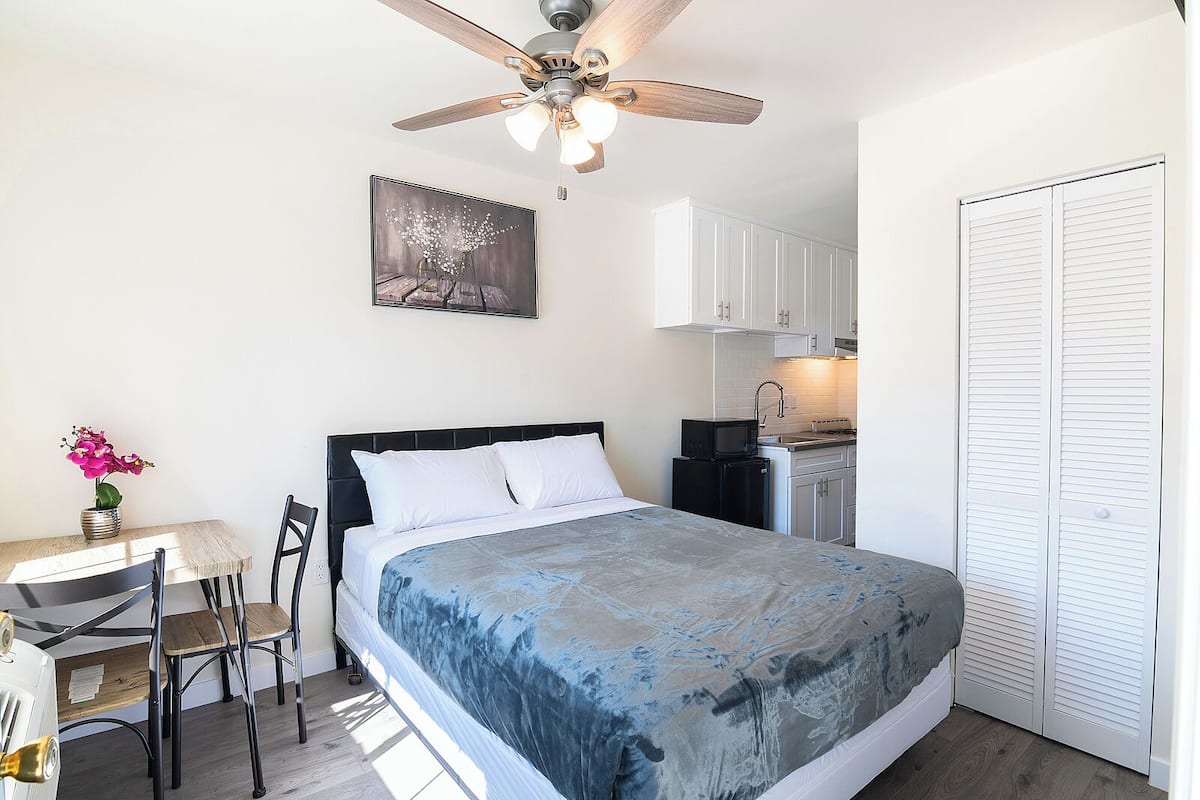 A bedroom with a bed, a desk, a chair, a ceiling fan, a microwave, and a white wardrobe.