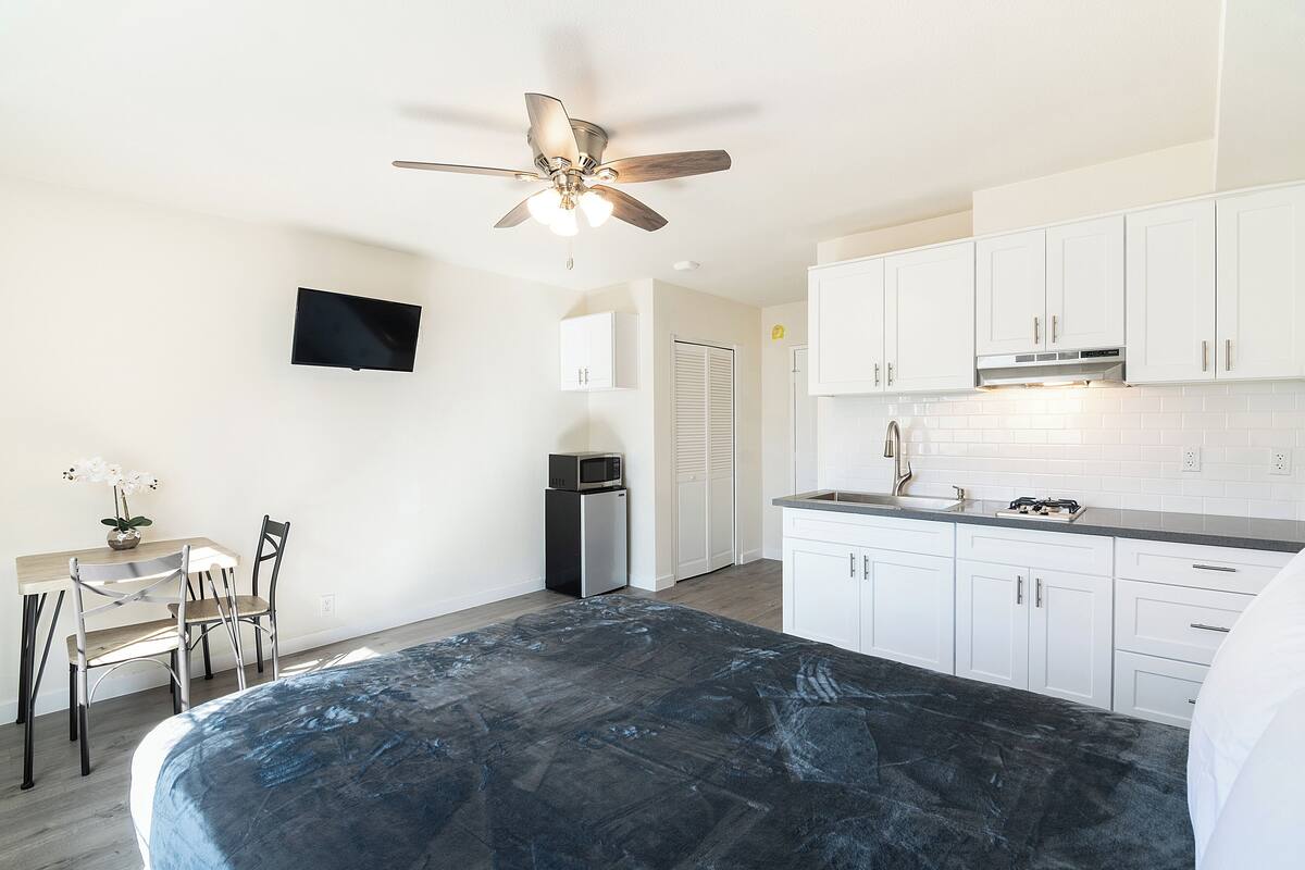 A bedroom with a bed, a desk with a chair, a television, and a kitchenette with cabinets and a microwave.