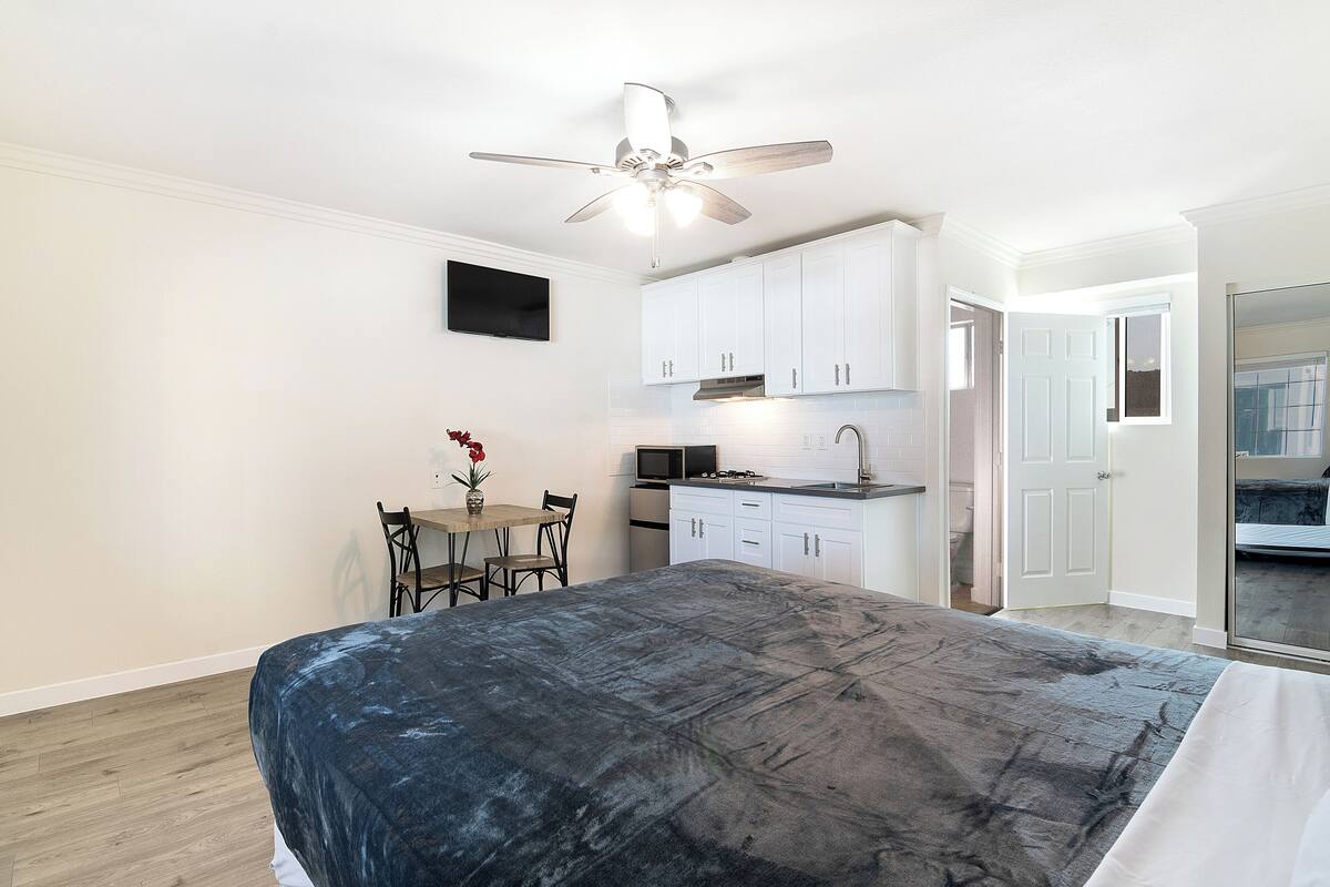 A bedroom with a bed, a small dining table with chairs, a ceiling fan, and a kitchenette with white cabinets and a microwave.