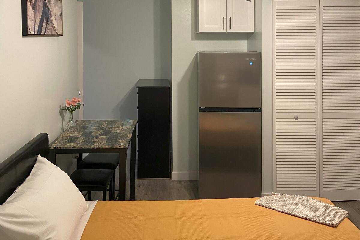 A compact room with a bed, a small table with a vase of flowers, a ceiling fan, a refrigerator, and a wardrobe.