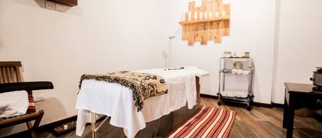 A room with a massage table, a wooden bench, a small shelf with items, and a patterned rug.