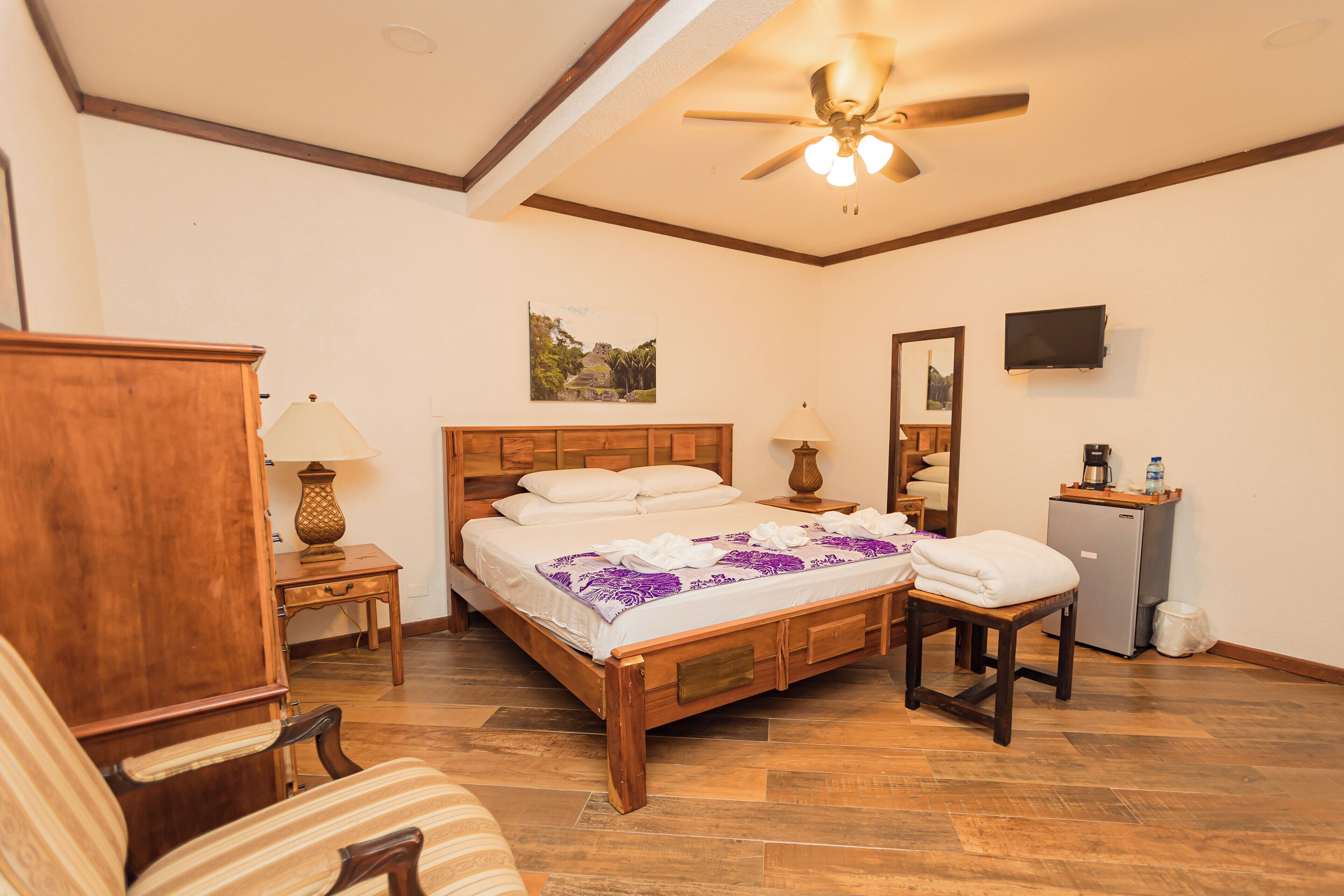 A bedroom with a wooden bed, a desk, a chair, a ceiling fan, a TV, and a refrigerator.