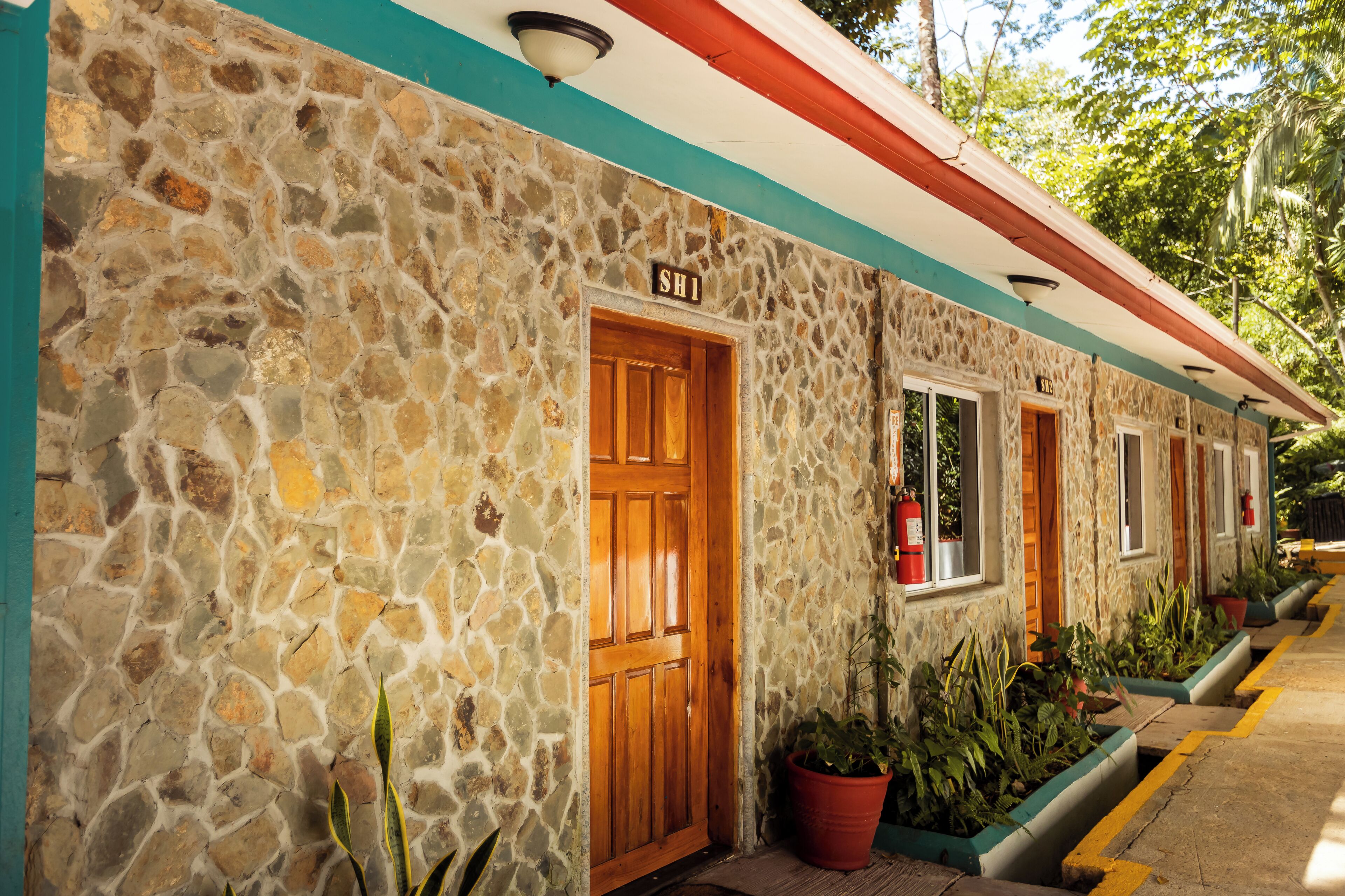 A stone building with a wooden door labeled "SH1," potted plants, and a red and white awning.