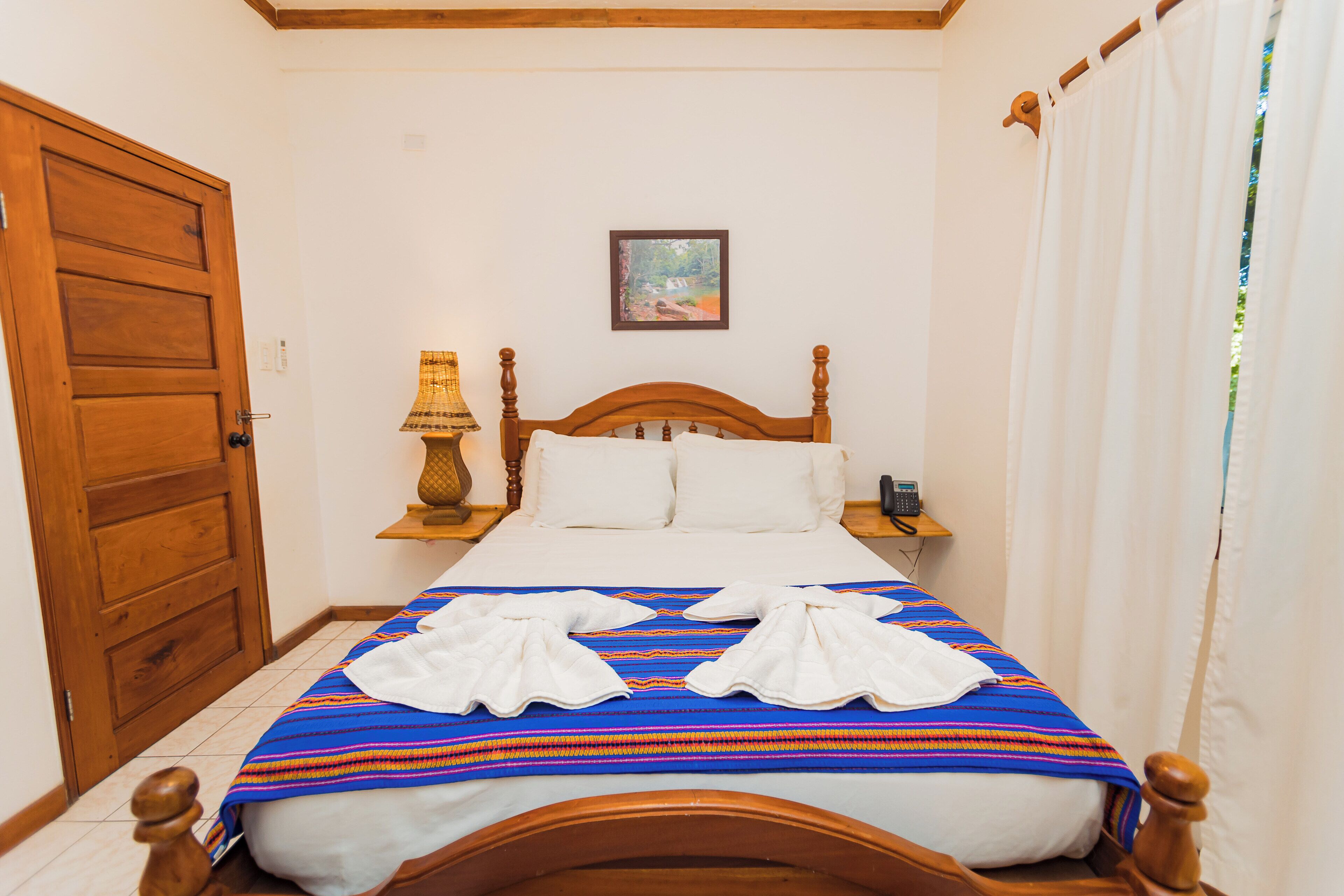 Una cama bien hecha con un edredón colorido, cabecera de madera y almohadas blancas. Se ve una puerta de madera a la izquierda y una pequeña mesa con una lámpara junto a la cama.