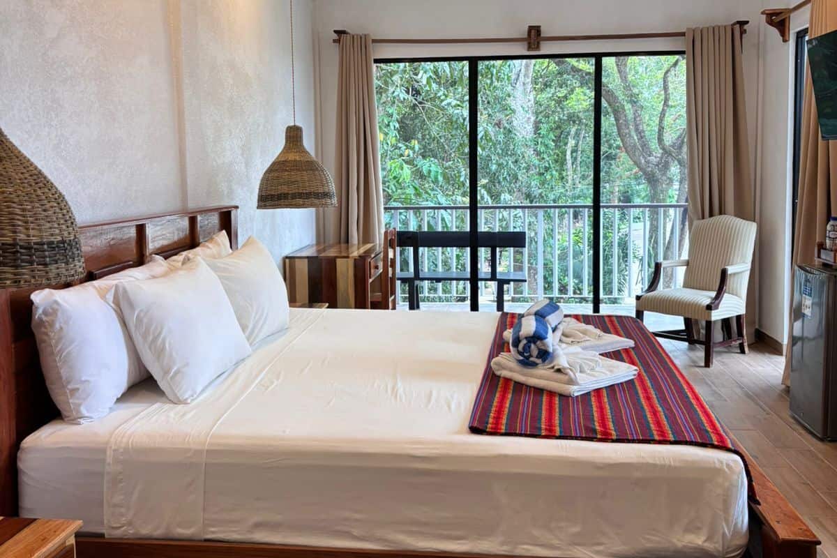Un dormitorio con una cama, ventilador de techo, ventana con cortinas, una silla y una mesita.