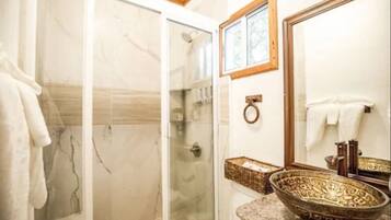 Un baño con ducha envuelta en vidrio, pared de azulejos de mármol, espejo con marco de madera y lavamanos decorativo.