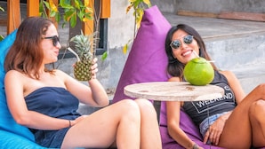 Two people sitting on a blue and purple couch, one holding a pineapple and the other holding a green drink with a straw. - Kaniu Capsule Hostel (Kuta)