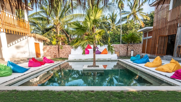 A swimming pool with colorful cushions, a palm tree, and a stone wall. - Kaniu Capsule Hostel (Kuta)