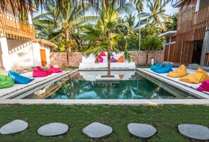 A swimming pool with colorful cushions, a palm tree, and a stone wall. - Kaniu Capsule Hostel (Kuta)