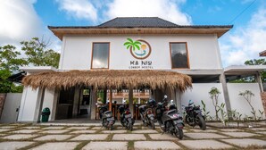 A building with a thatched roof entrance, a logo with a palm tree and a sun, and several motorcycles parked in front. - Kaniu Capsule Hostel (Kuta)