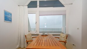 Zona pranzo con tavolo e sedie in legno, ampie finestre con persiane e vista sul mare.
