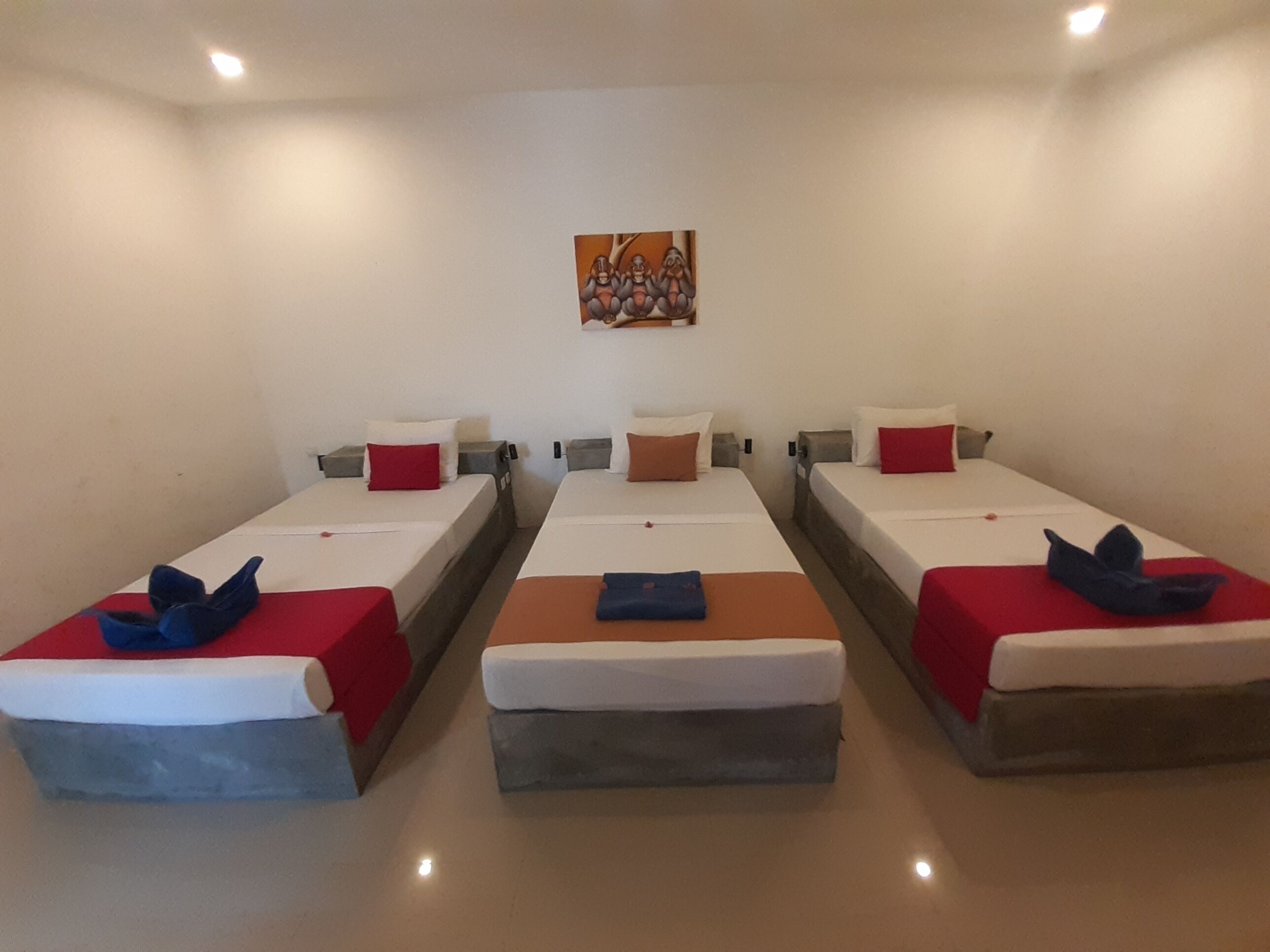 Een hotelkamer met drie eenpersoonsbedden, elk voorzien van een rood-witte spreidek, kussens en een blauw decoratief object op het bed.