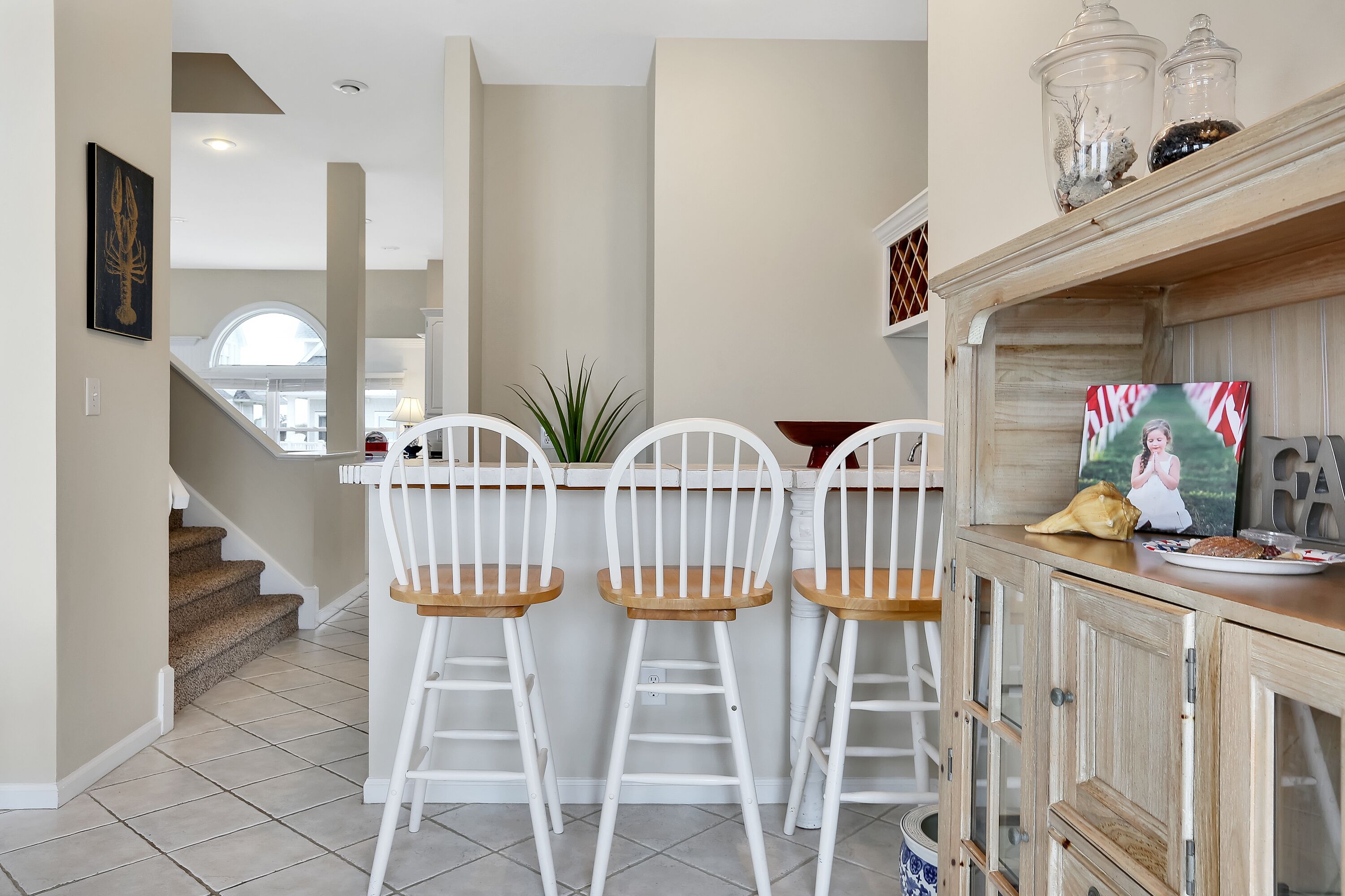 A kitchen with white bar stools, a wooden cabinet, and a staircase leading to an upper level.