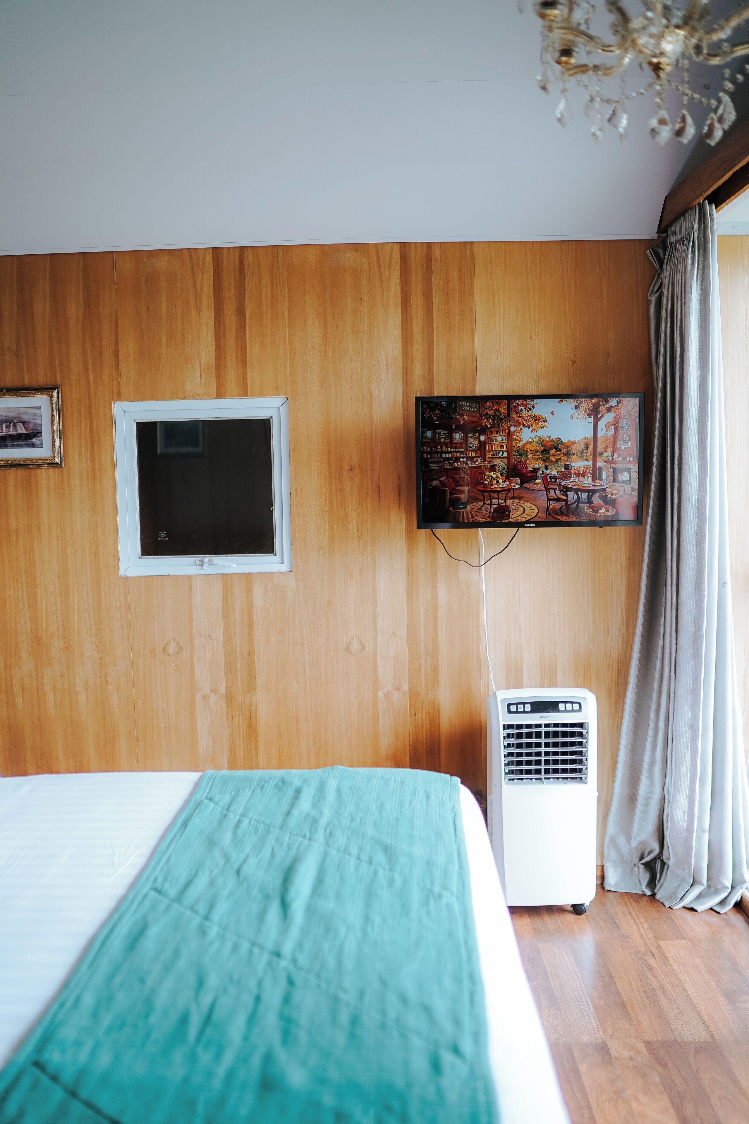 A hotel room with a wooden paneled wall, a television mounted above a window, and a standing air conditioner.