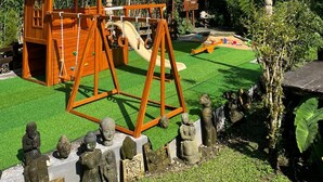 Una estructura de juego de madera con toboganes y columpios, rodeada de césped verde y estatuas de piedra.