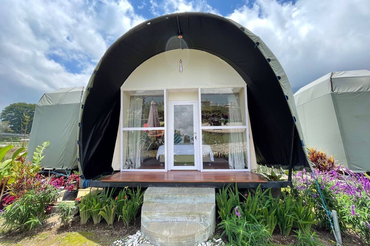 Una tienda en forma de cúpula con una terraza de madera y puertas de cristal, rodeada de exuberante vegetación de jardín.