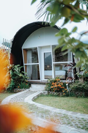 A house with a curved roof, a porch with a bench, and a garden with flowers and plants. - Pondanu Cabins By The Lake (Baturiti)