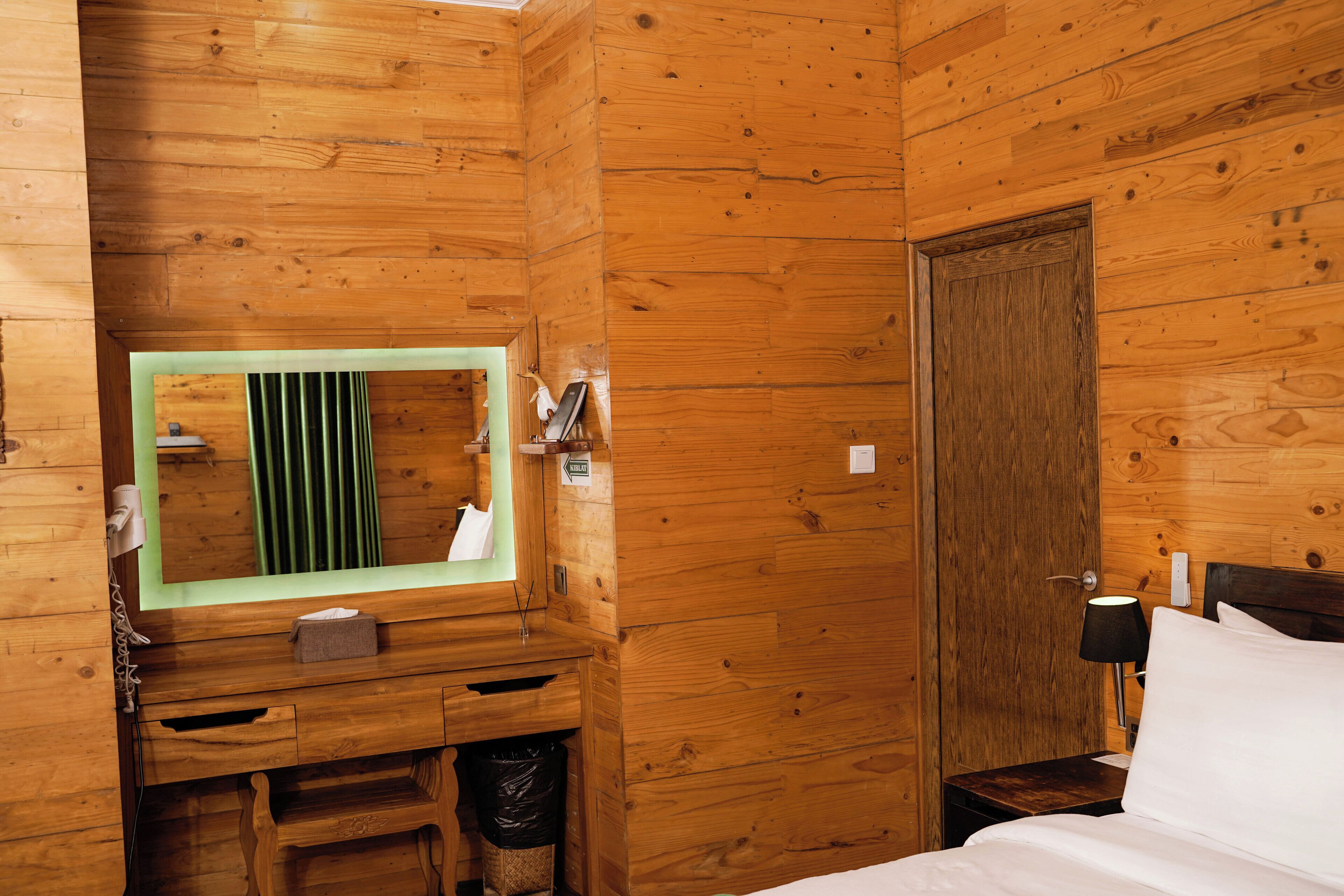 A wooden cabin room with a bed, a nightstand, a mirror, a small desk with drawers, and a trash bin.