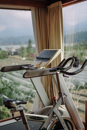An exercise bike with a digital display, positioned near a window with a view of greenery and mountains. - Pondanu Cabins By The Lake (Baturiti)