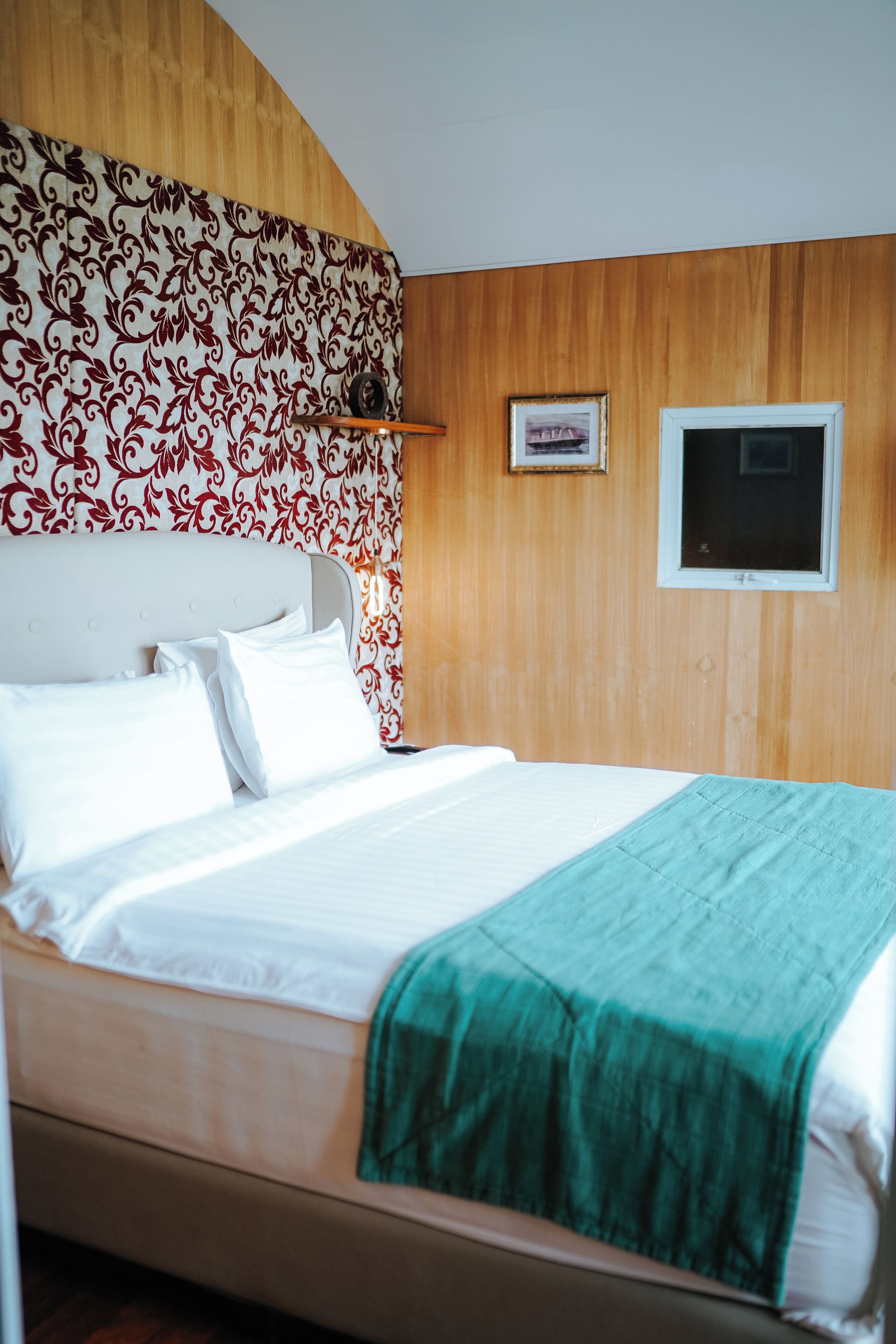 A neatly made bed with white linens and a green throw blanket, a wooden headboard with a floral pattern, and a small framed picture on the wall.