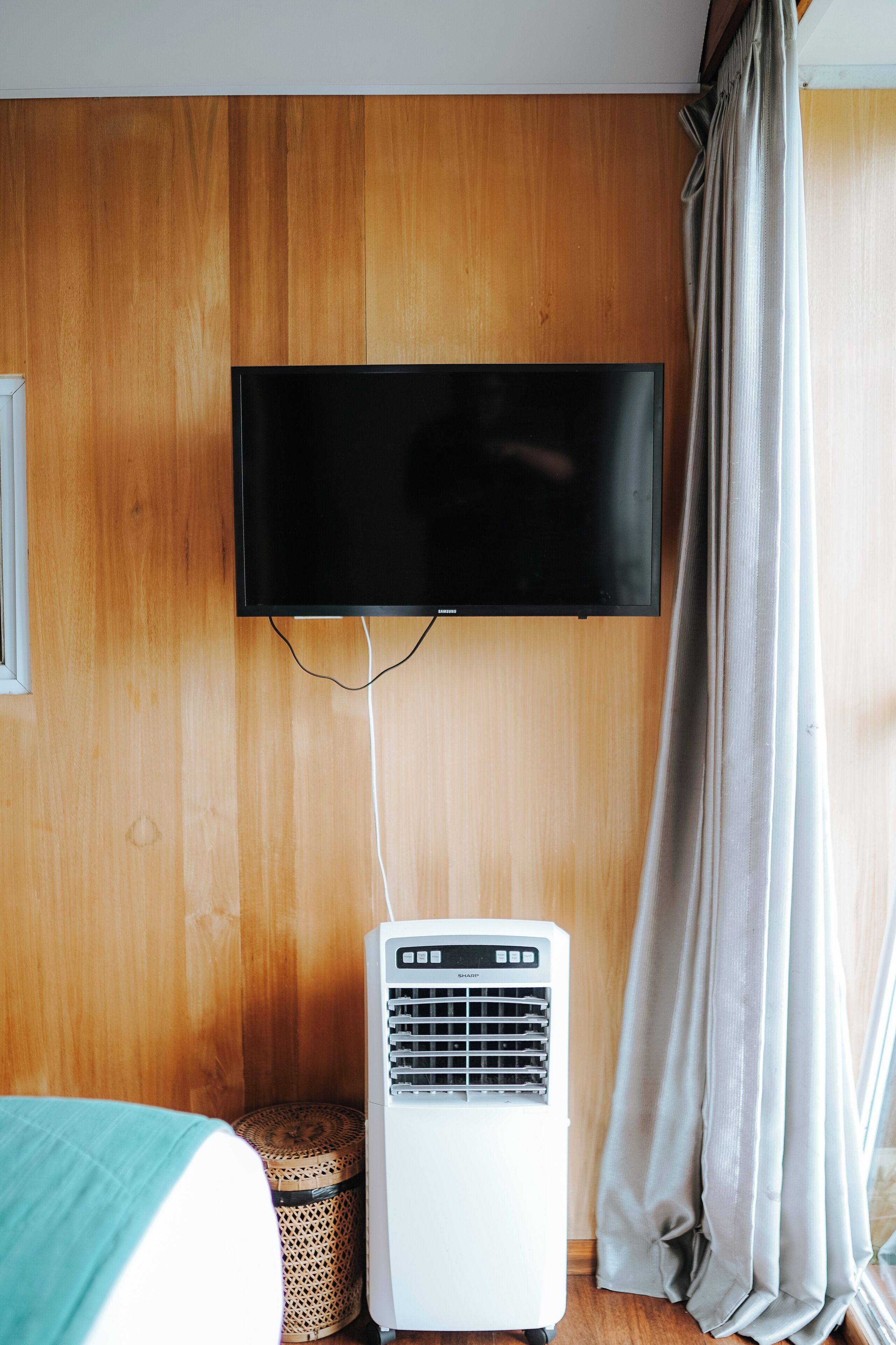 Una habitación con un televisor de pantalla plana montado, un aire acondicionado portátil blanco y una ventana con cortinas.