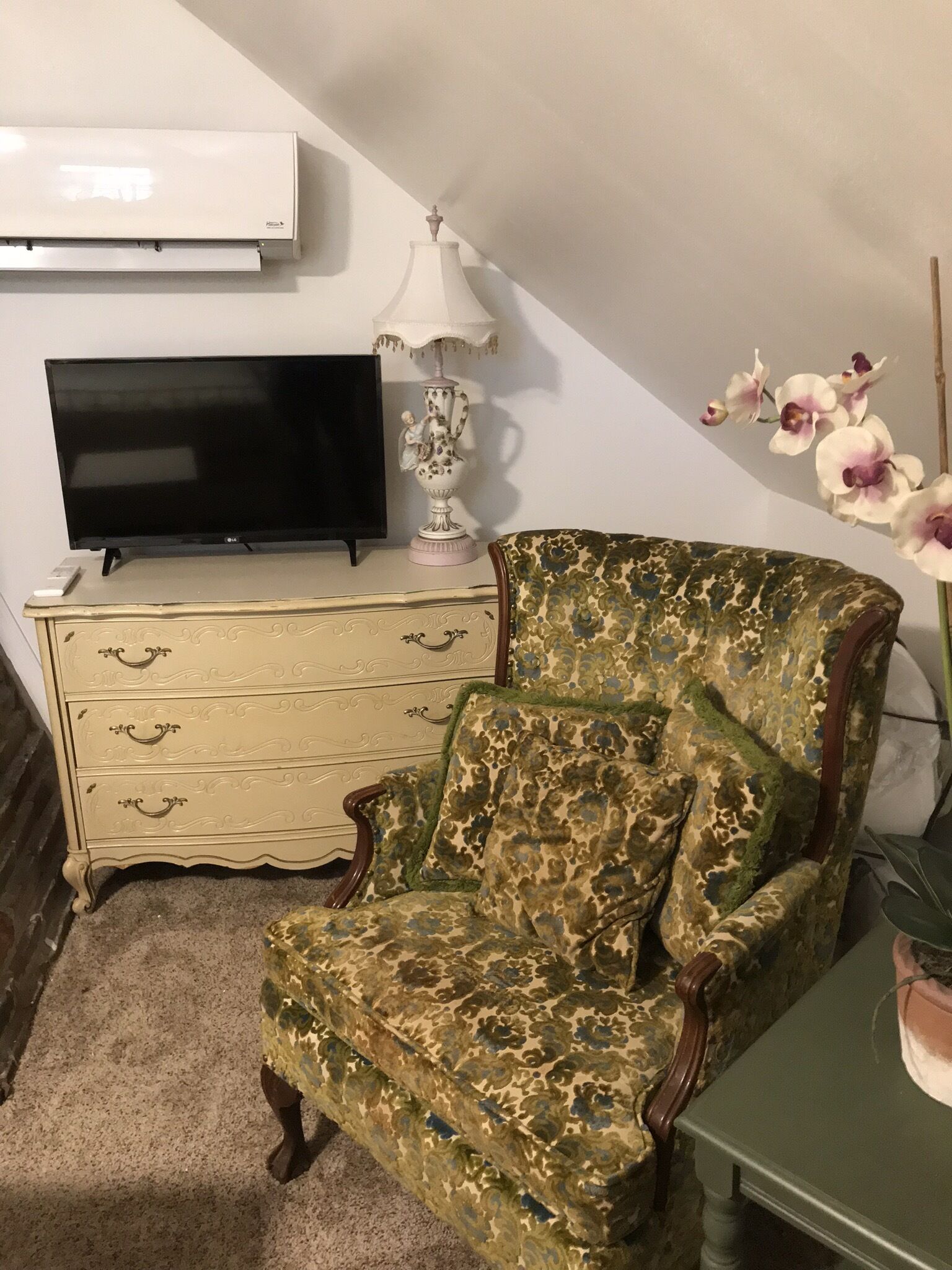 A room with a television on a white dresser, a floral-patterned armchair, and a potted orchid plant.
