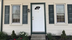 Une maison à deux étages avec une porte blanche, des volets verts et un petit porche agrémenté d’un paillasson.