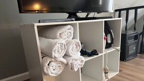 Valkoinen hylly, jonka päällä on rullalle taiteltuja pyyhkeitä, ja sen alla imuri sekä yläpuolella televisio.