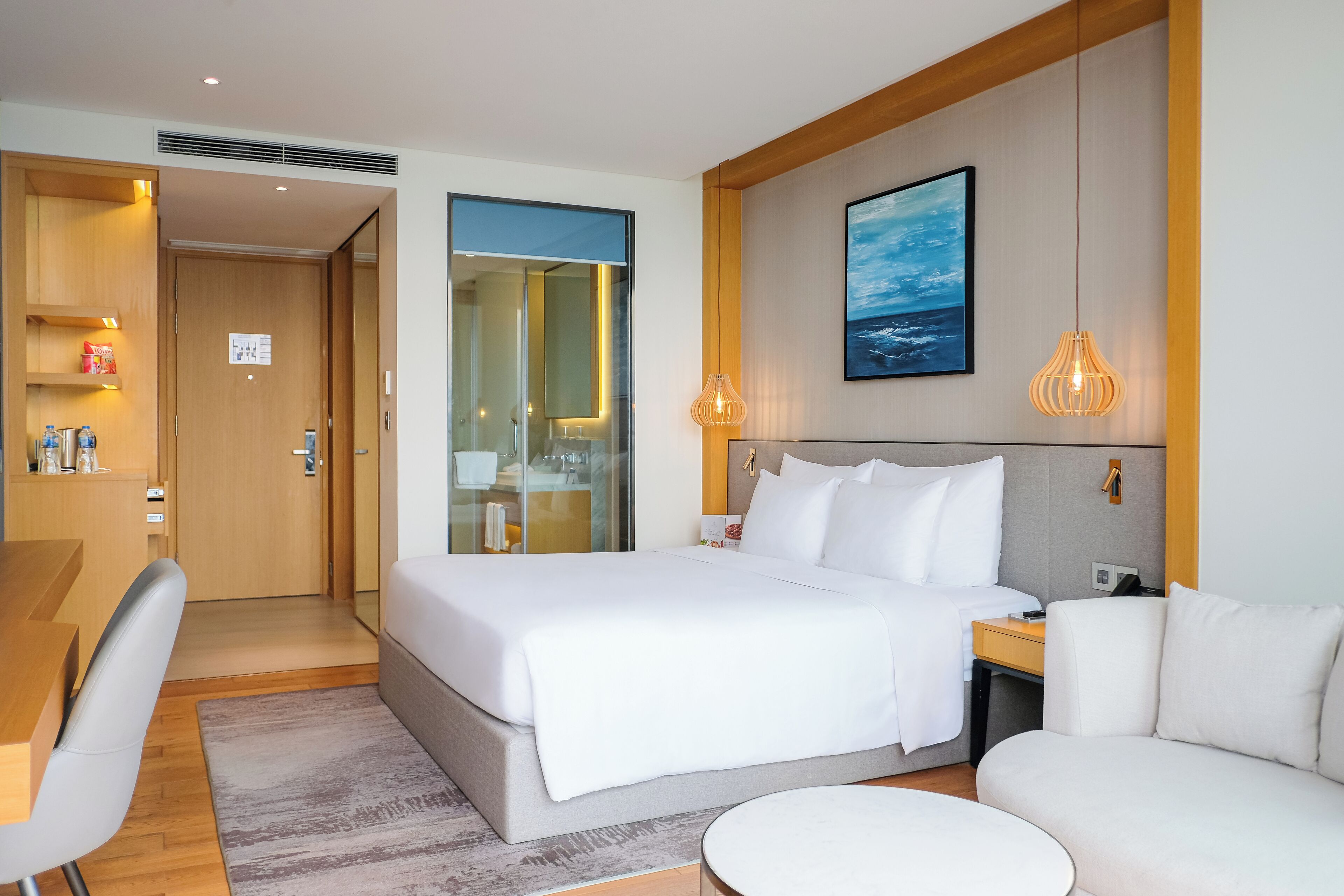 A modern hotel room with a large bed, a desk, a sofa, and a view of the sea through a glass door.