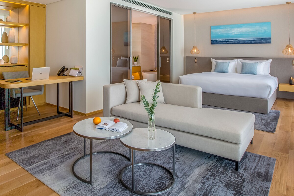 A modern hotel room with a bed, a sofa, a desk, and a small table with a plant and fruit.
