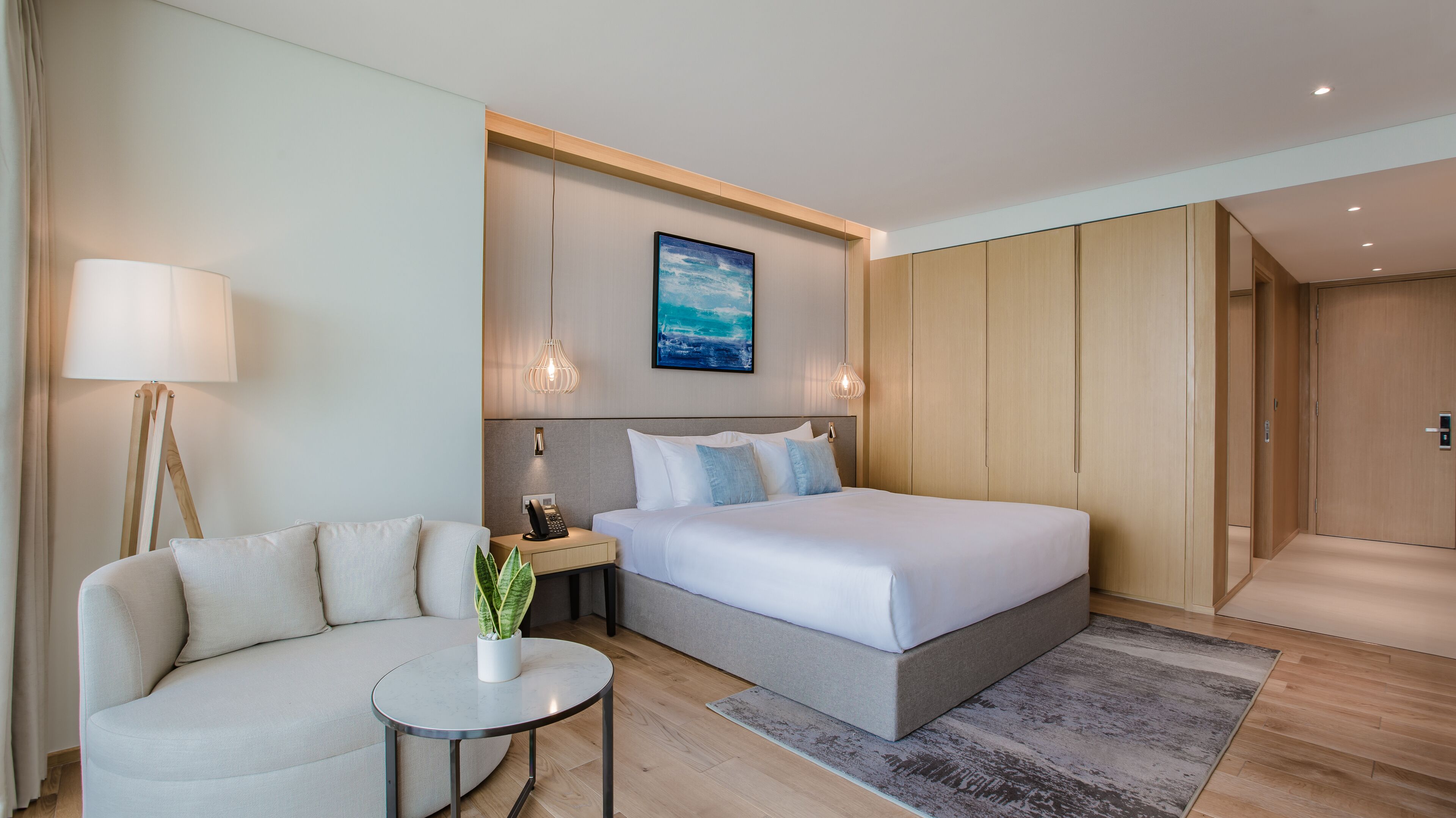 A modern hotel room with a bed, a sofa, a small table with a plant, a painting on the wall, and a wooden wardrobe.