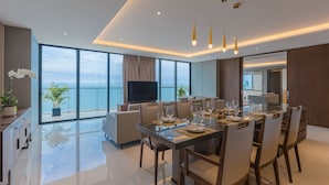 A modern living room with a large TV, comfortable seating, a dining area with a glass table, and a view of the sea.