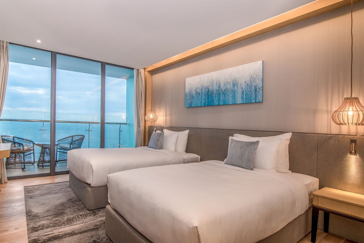 A modern hotel room with two beds, a large window with a view of the sea, a painting on the wall, and a small table with chairs.