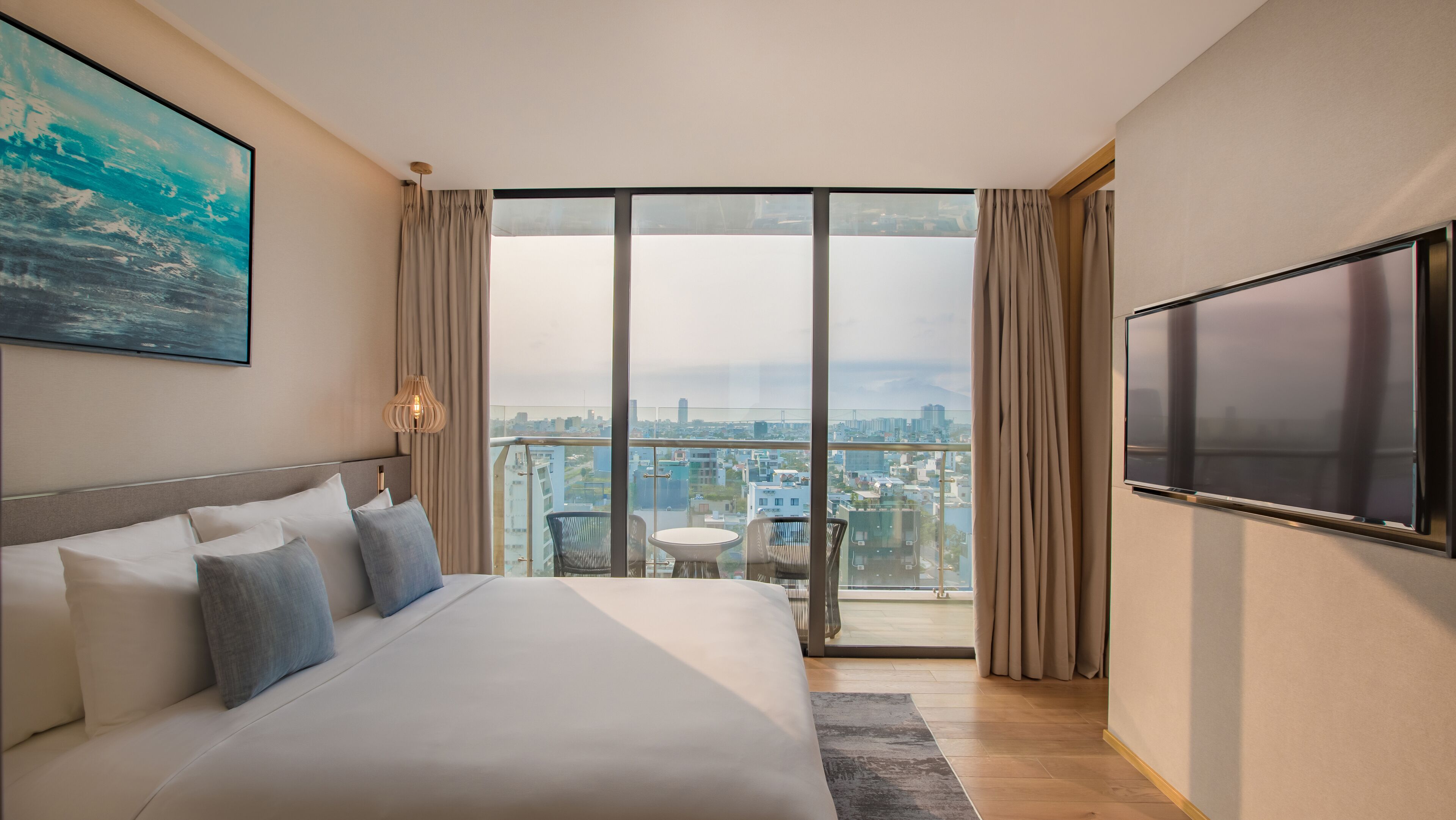 A hotel room with a large bed, a city view, a TV, and a balcony with a table and chairs.